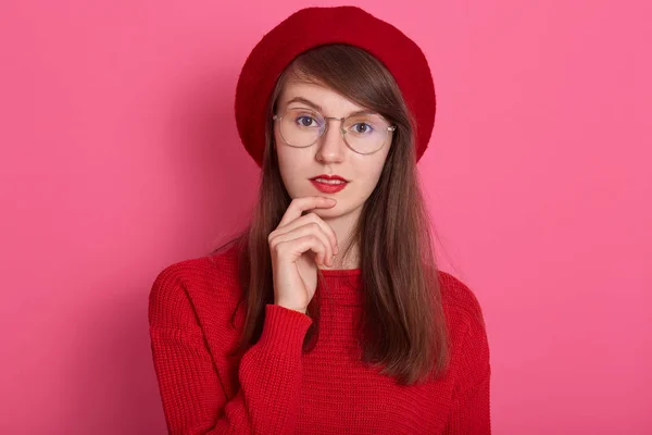 Close up retrato de mulher jovem mantém os dedos dianteiros perto do lábio inferior, estar em espírito elevado, vestindo boina vermelha, suéter e óculos, menina adorável em pé sobre a parede rosa, espaço de cópia para edvertisment . — Fotografia de Stock