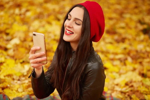 Egy kép aranyos lányról piros sapkában és fekete bőrdzsekiben az őszi parkban, amint önarcképet készít a mobilján. Vonzó hölgy pózol a szabadban. Emberek és technológiai koncepció. — Stock Fotó