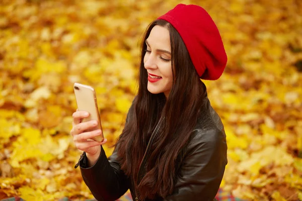 Az őszi parkban svájcisapkát és bőrdzsekit viselő gyönyörű lány szabadtéri képe önarcképet készít a mobilján. Lány gazdaság okostelefon, hogy fotó a szabad ég alatt a közösségi hálózatok. Az emberek fogalma — Stock Fotó