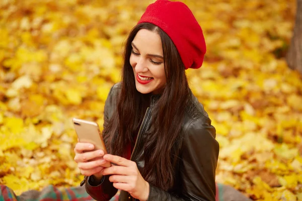 Image of beautiful Caucasian woman brunette dresses red beret and black leather jacket, walking through park, female holding smartphone and cheking social network, looks at device with smile. — Stock Photo, Image