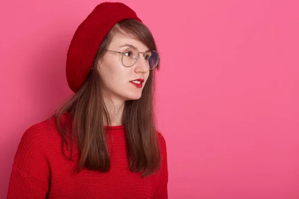 Close-up portret van pensive jonge vrouwelijke jurken trui, baret en ronde bril, op zoek bedachtzaam opzij, staande tegen een rooskleurige studio muur, kopieer ruimte voor reclame of promotie. — Stockfoto