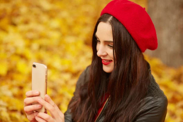 Portré fiatal divatos lány okostelefonnal a kezében, nézett devive, ül a földön a parkban ősszel. Trendy nő ruhák piros svájci sapka és kabát a parkban, selfie a szociális hálózat — Stock Fotó