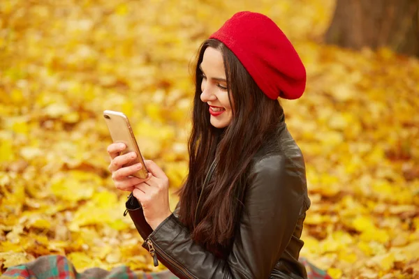 Image of attractive young girl with dark staright long hair, wearing red beret and leather jacket, looking at device screen with smile, model poses while sitting on ground covering yellow leaves. — Stock Photo, Image