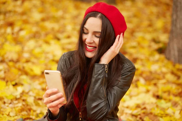 Emotional portrait of happy and relaxed beautiful young woman with dark hair, dresses beret and jacket, looks smiling at phone while sitting on fallen yellow leaves during walk in park. Autumn colors. — Stock Photo, Image
