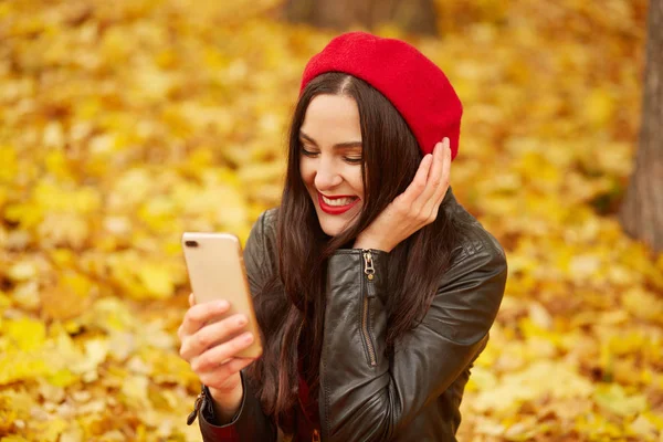 Close up outdoor portrait of woman, lady looks happy and taking self picture via smartphone in autumn park, young model posing on ground covering golden leaves. Technology and happiness concept.