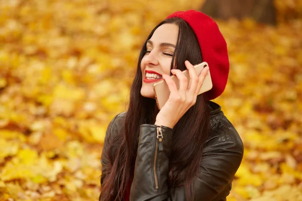 Внешний снимок молодой счастливой белой темноволосой женщины со смартфоном, очаровательная дама, сидящая на земле, окруженная желтыми листьями, в кожаной куртке и красном берете. Люди и технологии . — стоковое фото