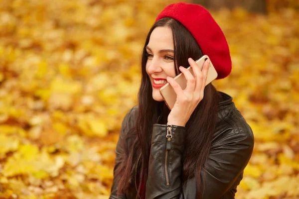 Горизонтальный снимок веселой модной женщины в кожаной куртке и красном берете осенью. Красивая юная брюнетка, держит смартфон, разговаривает с парнем. Золотой осенний фон — стоковое фото