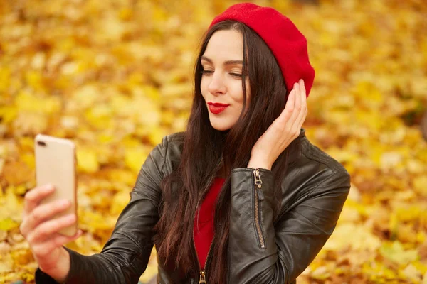 Portré vidám fiatal nő körül őszi levelek, pózol őszi parkban tele lombozat, hogy szelfi az ő okostelefon, ruhák piros sapka és fekete bőrkabát. Az emberek fogalma. — Stock Fotó