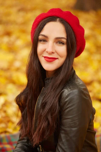 Mooie vrouw in het herfstpark, genietend van warm zonnig weer. Close-up beeld van vrolijke donkerharige jonge dragen zwart lederen jas en rode baret, op zoek met charmante glimlach direct op de camera. — Stockfoto