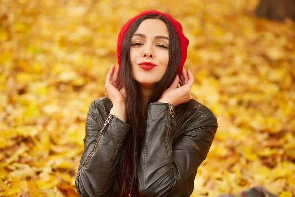 Горизонтальный снимок красивой женщины, показывающей жест поцелуя и улыбающейся в камеру, позирующей на фоне осеннего парка, очаровательная девушка одевается в красный берет и кожаную куртку, окруженная желтыми листьями . — стоковое фото