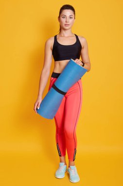 Spor kıyafetleri giyen, ayakta duran ve elinde mavi spor minderiyle kameraya bakan, yoga seansı veya antrenmandan sonra formda olan genç, güzel, sarı saçlı bir bayan sporcu. Spor konsepti.