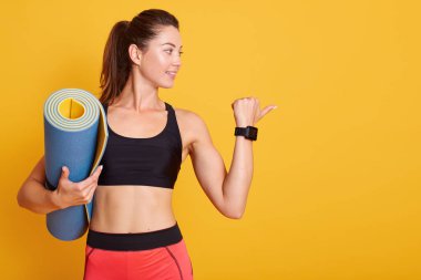 Güçlü güçlü beyaz kadın kolunu kaldırıyor, pazılarını gösteriyor, beden eğitimi için mavi spor minderi tutuyor, spor giysileri giyiyor, sarı arka planda izole edilmiş, çalışmaya hazır bayan ya da yoga antrenmanına hazır..