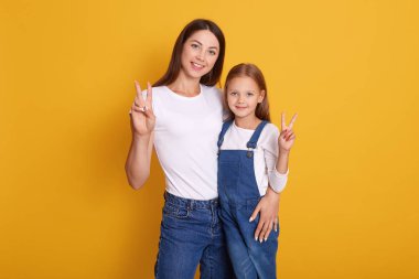 Sevimli kızıyla barış işareti gösteren mutlu bir kadının portresi, beyaz tişört ve kot giyen bir kadın, çocuk mercan giyiniyor, mutluluğunu ifade ediyor, sarı stüdyo arka planında izole edilmiş..