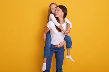 Stüdyo çekimlerinde güzel esmer anne beyaz gömlekli, tulumlu, kadın elbiseli ve kot pantolonlu, sarı arka planda yalnız poz veren küçük sarışın kızını kucağına alır..