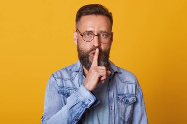 Ciddi sakallı bir adamın işaret parmağını dudaklarının yakınında tutarken, sarı arka planda izole bir şekilde poz verdiği yatay bir görüntü. Stüdyoda duran yakışıklı adam şık bir kot ceket giyiyor. Halk konsepti.