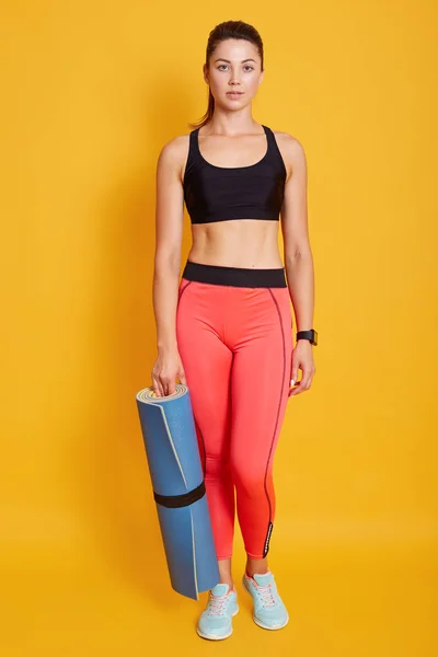 Full length πορτρέτο της νεαρής γυναίκας με μπλε στρώμα γυμναστικής στο χέρι, θέτοντας απομονωμένη πάνω από κίτρινο φόντο, κοιτάζοντας directlky στην κάμερα, έτοιμο για αθλητικές ασκήσεις, γυναικεία φορέματα κομψό αθλητικό ένδυμα. — Φωτογραφία Αρχείου