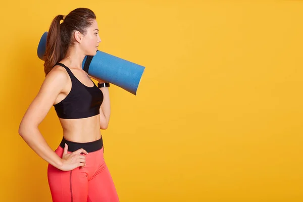 Côté wiev de jeune belle femme caucasienne debout et tenant tapis de yoga bleu sur l'épaule, étant prêt pour l'exercice dans la salle de gym, portant des vêtements de sport élégants. Espace de copie pour la publicité. Concept sportif . — Photo