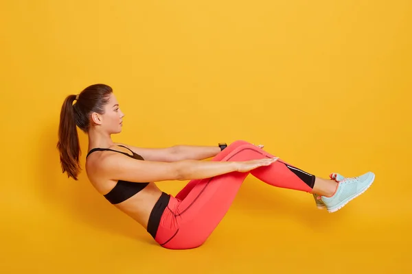 Indoor shot of young attractive sporty woman practicando fitness, haciendo ejercicio en barco, ejercitándose, usando sujetador negro y leggins rojos, longitud completa, aislado sobre fondo de estudio amarillo, vista lateral . — Foto de Stock