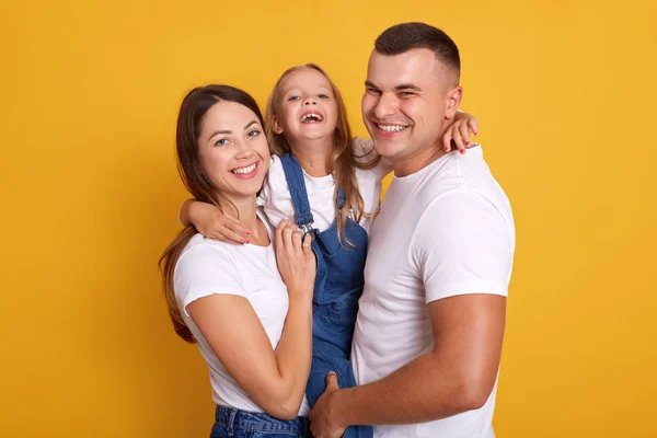 Plan horizontal d'une famille heureuse avec un enfant, souriant directement à la caméra, posant isolé sur fond de studio jaune, portant des t-shirts blancs décontractés. Concept de convivialité et de bonheur . — Photo