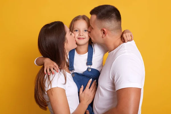 Εσωτερική λήψη χαρούμενης νεαρής οικογένειας με μια μικρή κόρη να ποζάρει μαζί απομονωμένη σε κίτρινο φόντο, γονείς να φιλούν την κόρη τους, χαριτωμένο ξανθό παιδί να κοιτάζει την κάμερα. Έννοια της στεγανότητας. — Φωτογραφία Αρχείου