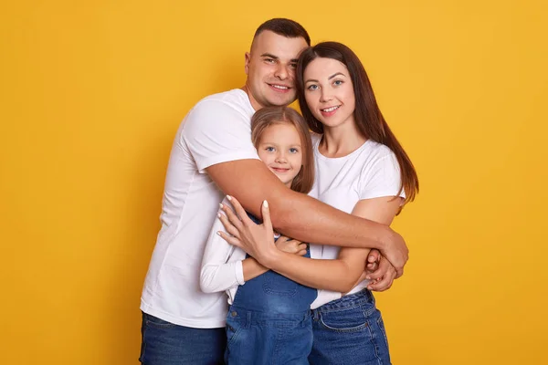 Horizontal shot of beautiful young family hugging, looking directly at camera and smiling while standing against yellow studio wall, people dress casually, expressing happyness. Relationship concept — Stock Photo, Image