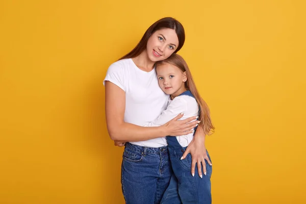 Κοντινό πορτραίτο της όμορφης νεαρής μητέρας και της γοητευτικής μικρής κόρης της, στέκεται χαμογελώντας στον κίτρινο τοίχο του στούντιο, γυναίκα αγκαλιάζει το παιδί της με μεγάλη αγάπη, φορώντας λευκό πουκάμισο και denim τρούνες — Φωτογραφία Αρχείου