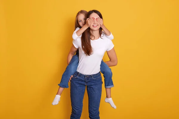 Studio shot of woman giving pyggyback ride indoor, female wearing white casual t shirt and jeans, charming kid dresses overalls closing her mommy's eyes, isolated on yellow background. Family concept. — 스톡 사진