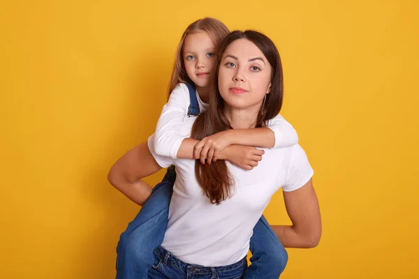 Close up portrait of mother giving piggyback ride to her cute daughter and being in good mood, models posing isolated over yellow background, woamn wearing white t shirt holding her blonde kid. — 스톡 사진