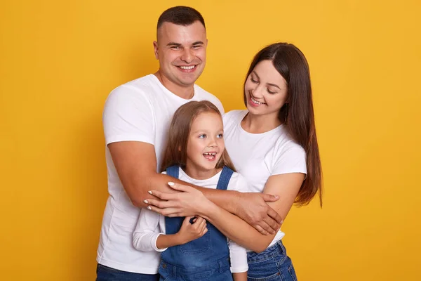 Happy family looking very excited, posing isolated over yellow background, mother, father and daughter wearing white shirts, woman and man hugging charming girl with great love. Happyness concept. — Stock Photo, Image