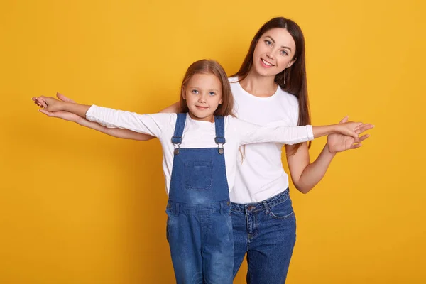Woman dresse casual clothes having faun with her cute child baby girl wearing white shirt and denim overalls. Mother, little kid posing isolated over yellow backround. Family and childhood concept. — 스톡 사진