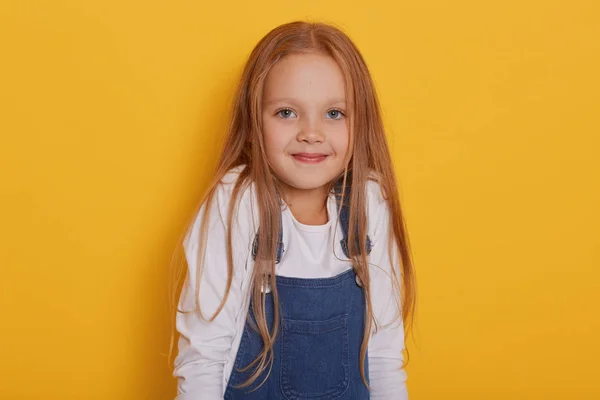 Tiro horizontal de niña linda pelo rubio, vestidos de camisa blanca y overoles de mezclilla, se ve tímido, posando aislado sobre el fondo amarillo, niño lindo apretando hombros, se levanta sonriendo . — Foto de Stock