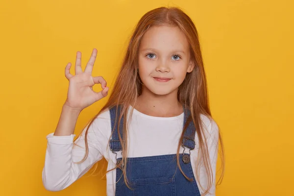 Ritratto ravvicinato di giovane ragazza bionda di sette anni che indossa camicia bianca a maniche lunghe e tuta jeans, modello in posa isolata su sfondo giallo, mostrando segno ok, guardando direttamente la fotocamera . — Foto Stock