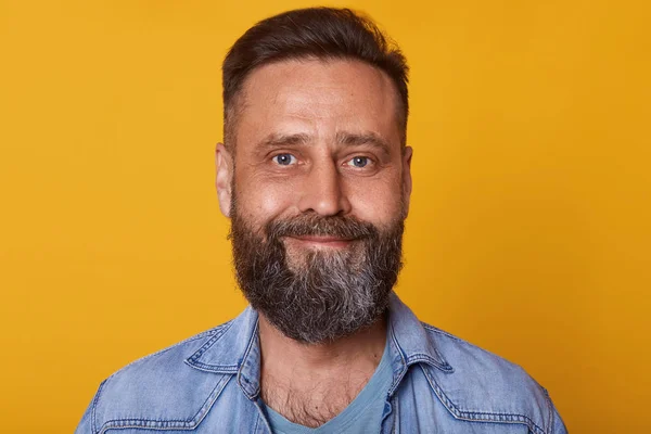 Portrait of single bearded handsome young Caucasian man with cute smile,male wearing denim jacket, posing isolated over yellow background, looking smiling directly at camera. People concept. — Stock Photo, Image