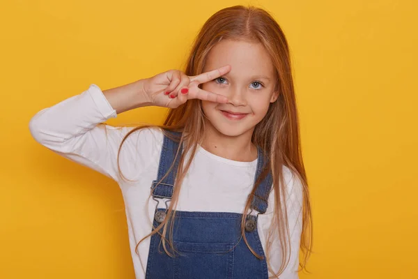 Close up ritratto di bella attraente bella allegra ragazza bionda dai capelli dritti mostrando segno v vicino al suo occhio isolato su sfondo giallo vivo luminoso, bambino veste camicia bianca. Lingua dody . — Foto Stock