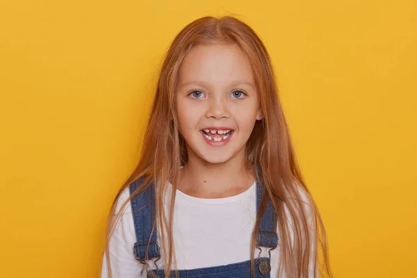 Cheerful child without front teeth posing with opened mouth and funny smile. Nice little blonde girl with milk teeth fell out, stands against yellow wall, dresses denim overalls. Childhood concept. — Stock Photo, Image
