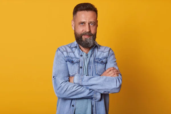 Representative, personable man with beard looking directly at camera, stands with arms folded, isolated over yellow background, dresses stylish denim jacket, having seriousfacial expression. — Stock Photo, Image