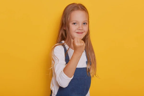 Ritratto di ragazza emotiva isolata su sfondo giallo, grazioso bambino biondo che mostra il pugno, bambino che indossa camicia bianca e ovealls, essendo arrabbiato, guardando direttamente la fotocamera. Concetto di infanzia . — Foto Stock