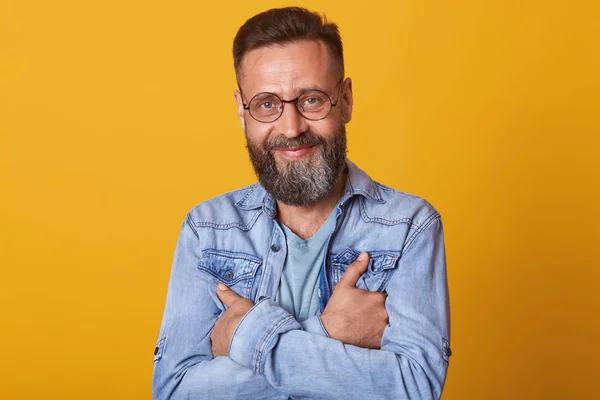 Close up portrait of handsome young man looking directly at camera through his glasses and smiling while standing against yellow studio background, dresses stylish denim jacket, looks happy. — Stock Photo, Image