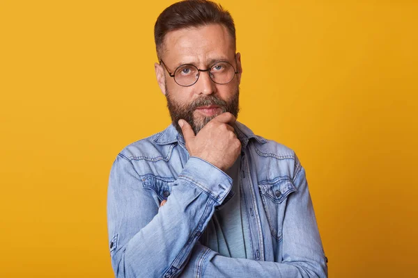 Studio shot of pensive male touching his perfect beard. Close up portrait of middle aged handsome bearded man standing against yellow background, attractive man wearing denim jacket. People concept. — Stock Photo, Image