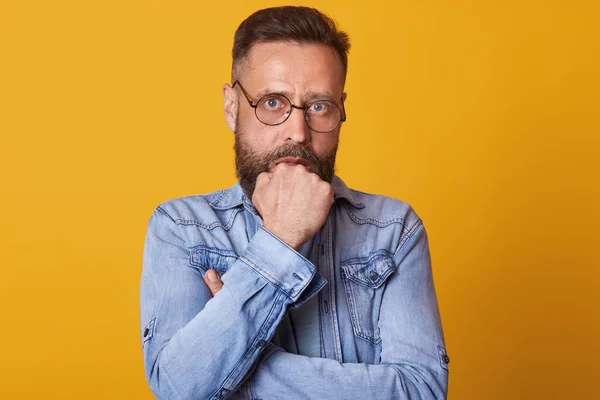 Portrait of amazed Caucasian male holds hands in fists under chin, looks worried, has eyes popped out, being frightened by unexpected news, dressed denim jacket, posing isolated over yellow background — Stock Photo, Image