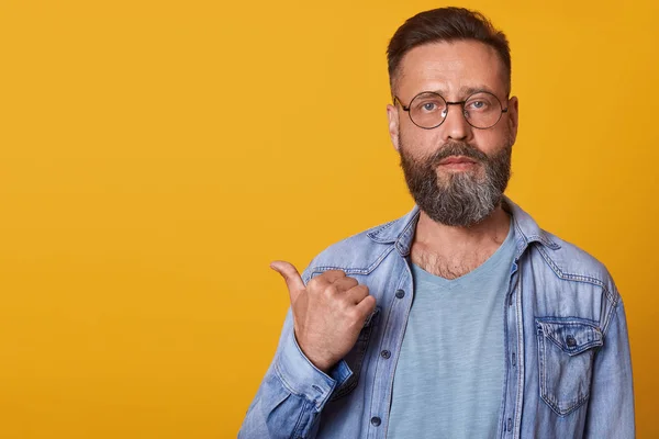 Photo of positive bearded man wearing casual gray t shirt, denim jacket and spectacles. Male advertising product with pointing finger aside on copyspace isolated over yellow studio background. — Stock Photo, Image