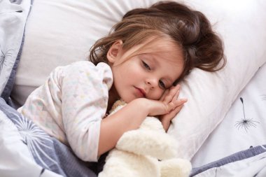 Beyaz yumuşak köpekli, yatakta dinlenen, oyuncağını kucaklayan, sıcak battaniyenin altında gözlerini açık tutan, siyah saçlı, beyaz pijama giyen çekici bir bayan çocuk. Sabah konsepti.