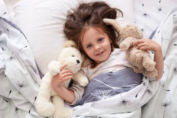 Image of girl lying with fluffy teddy bear and dog before giving them big hug, beautiful kid relaxing in bed with her toy, dark haired child wearing white pajama, charming kid expressing happyness. — Stock Photo, Image