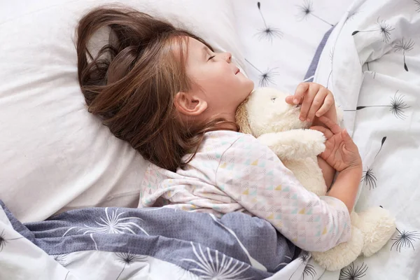 Cute little girl laying in bed, fall asleep after whole day playing, lying on white pillow under blanket with closed eyes, charming kid wears pajama, female kid relaxing in her room. Childhood concept — Stock Photo, Image