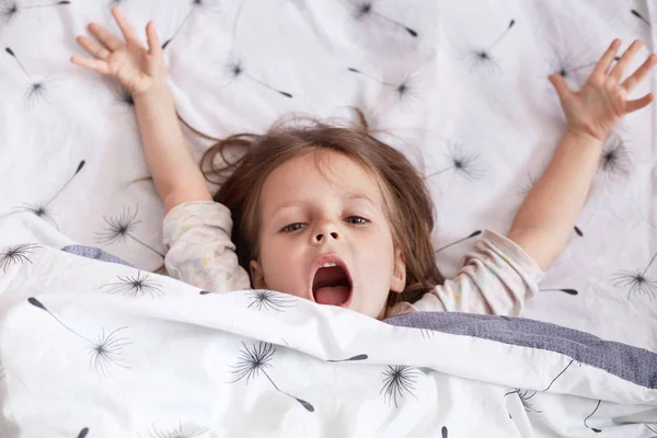 Indoor of cheerful little girl lying in bed under blanket with dandelion, charming kid wearing pajama, keeps hands up and mouth opened, screams something happily. Morning and childhood concept. — Stock Photo, Image