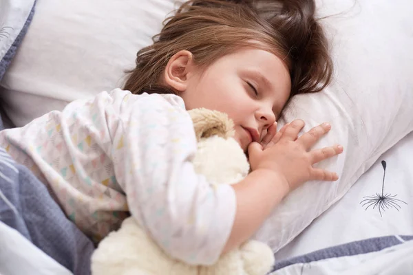 Indoor shot of pretty little girl hugging white soft dog toy while sleeping in bed at home, child spending morning time in cosy room, keeps eyes closed, having dark hair, desses white pajama. — Stock Photo, Image