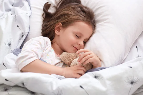 Close up portrait of beautiful girl sleeping in pajama in bed with her teddy bear, lying on pillow with closed eyes, charming cute female kid having dark hair. Childhood and morning time concept. — Stock Photo, Image