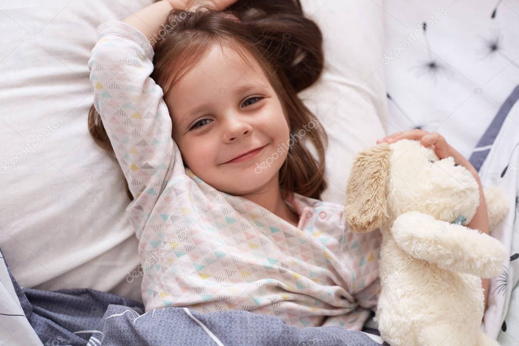 Top view of cute awaking girl with her toy animal lying on pillow under blanket, looking smiling directly at camera, charming female kid expressing happyness. Happy family and childhood concept.