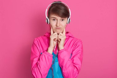 Kafasında beyaz kulaklık olan, müzik dinleyen, üzgün görünen, parmaklarını yanağında tutan beyaz spor giysili neşeli beyaz bir kızın stüdyo fotoğrafı. Sağlıklı yaşam tarzı, zindelik, sağlık konsepti.