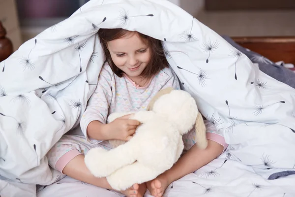 Portrait of cute girl and dog toy under blanket, little female kid and dog toy playing together games under white blanket, chirming child spending morning time in cosy room. Childhood concept. — Stock Photo, Image